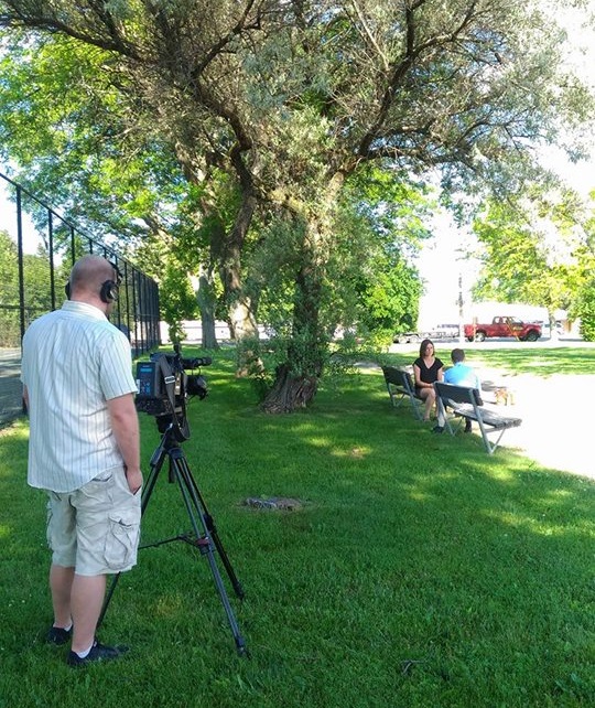 Photo shows a tv camera rolling in the foreground left. The camera is pointed at two women who sit on a park bench on the right side of the photo. They are talking, and clearly being recorded by the camera.