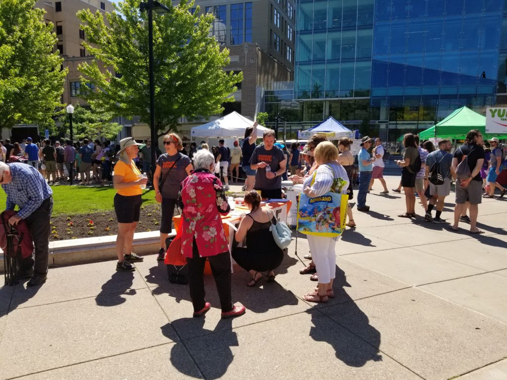 Many people are milling around tents and tables outside. A crowd of people surrounds an orange table with WAVE information, and two people in WAVE shirts are talking with the people gathered.