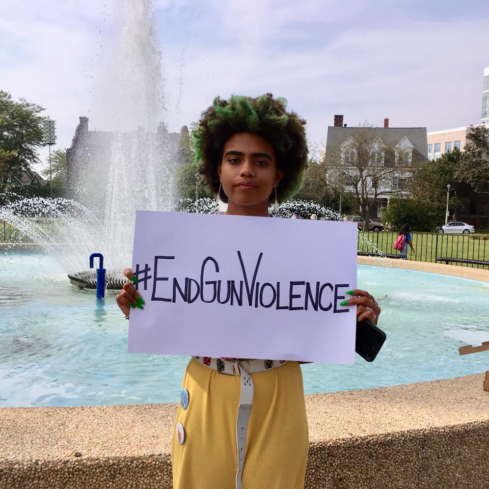 Young person standing by a fountain holding a sign that says "#End Gun Violence"