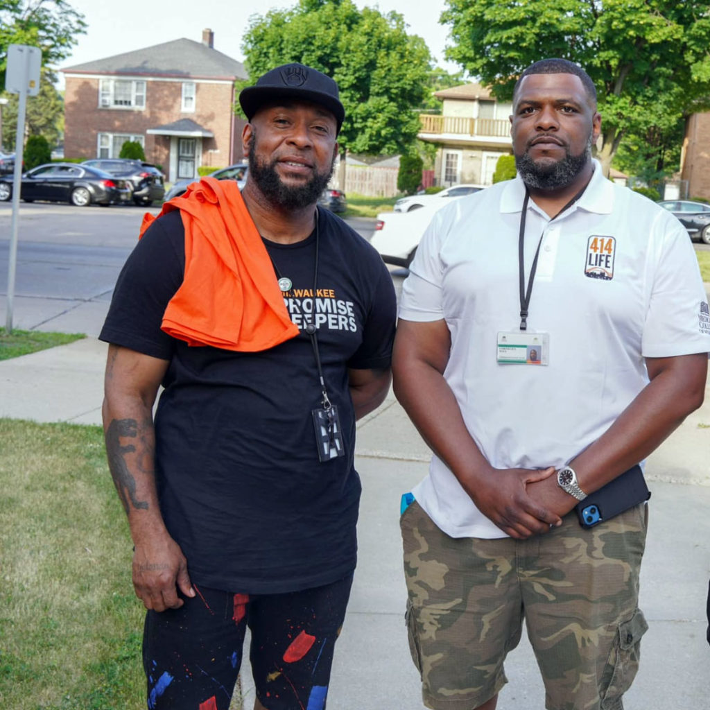 Two men stand facing the camera. One wears a shirt that says "Promise Keepers"; the other has a shirt that says "414Life." Both of these programs are violence intervention programs in Milwaukee.