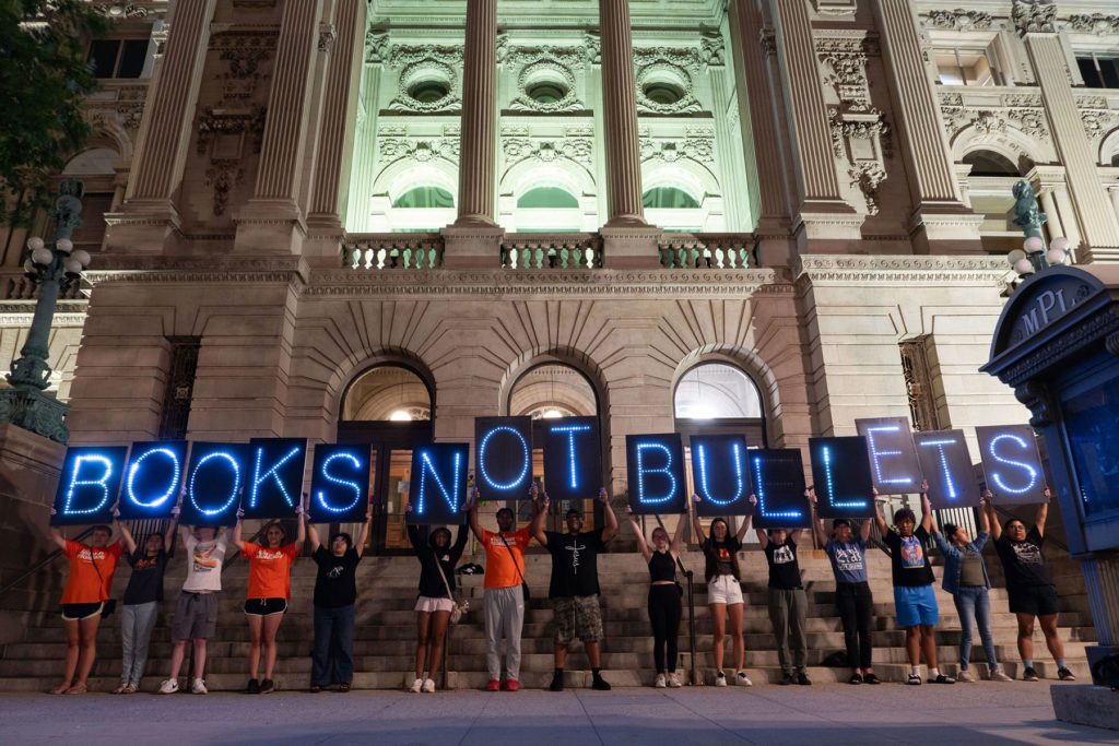 Photo courtesy of Overpass Light Brigade. A line of people stand holding light-up letters that spell out books not bullets.