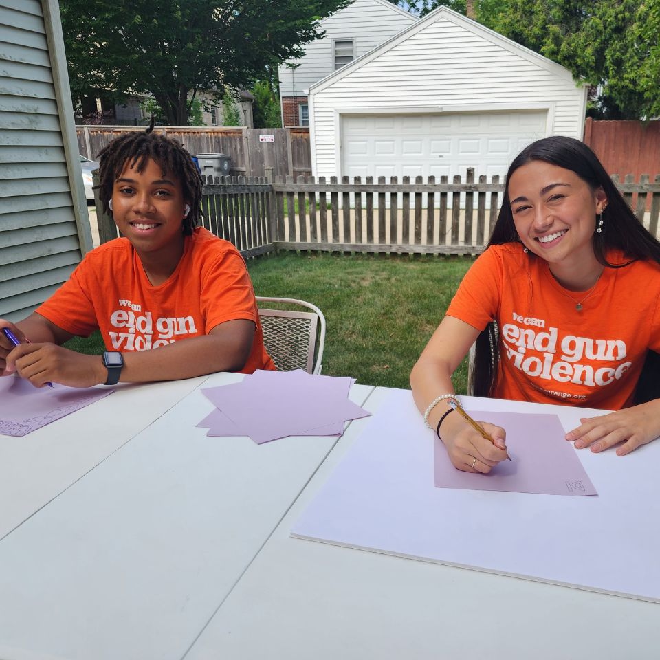 Two teens sit at a table outside and smile at the camera. They are both holding pencils, and have poster board. They appear to be making posters.
