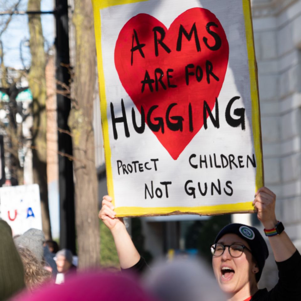A person at a rally holds a sign above their head. The sign says "Arms are for hugging."