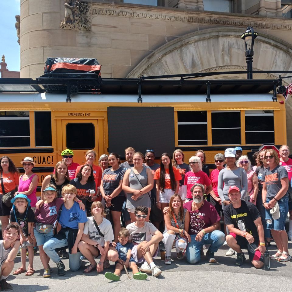 Several rows of people crowd into a photo frame for a picture in front of a yellow school bus. Participants range in age from 5 or 6 to elderly.