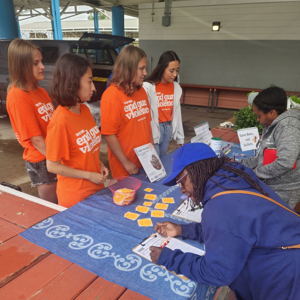 Four teens in orange "end gun violence" shirts stand behind a table. The table is set up with stickers and information sheets to educate people about gun violence. Two people on the other side of the table are signing up to receive information.