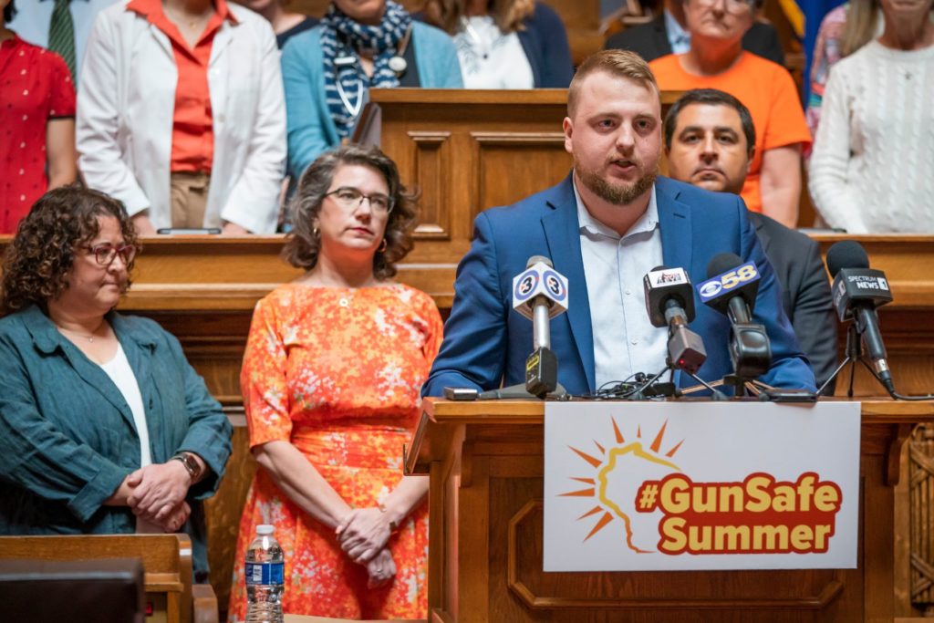 WAVE's policy manager stands at a podicum at a press converence. The podium has a sign that says #GunSafeSummer. Wisconsin Attorney General Josh Kaul and several state legislators stand behind, listening.