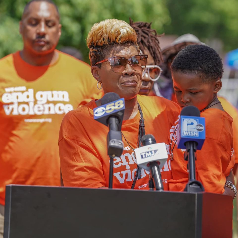 A woman wearing an orange "end gun violence" shirt speaks at a podium with microphones posted on it. She is holding back tears, and holding a young boy in her arms, of about 4 years old. Behind her other people stand, also wearing orange shirts that say "end gun violence."