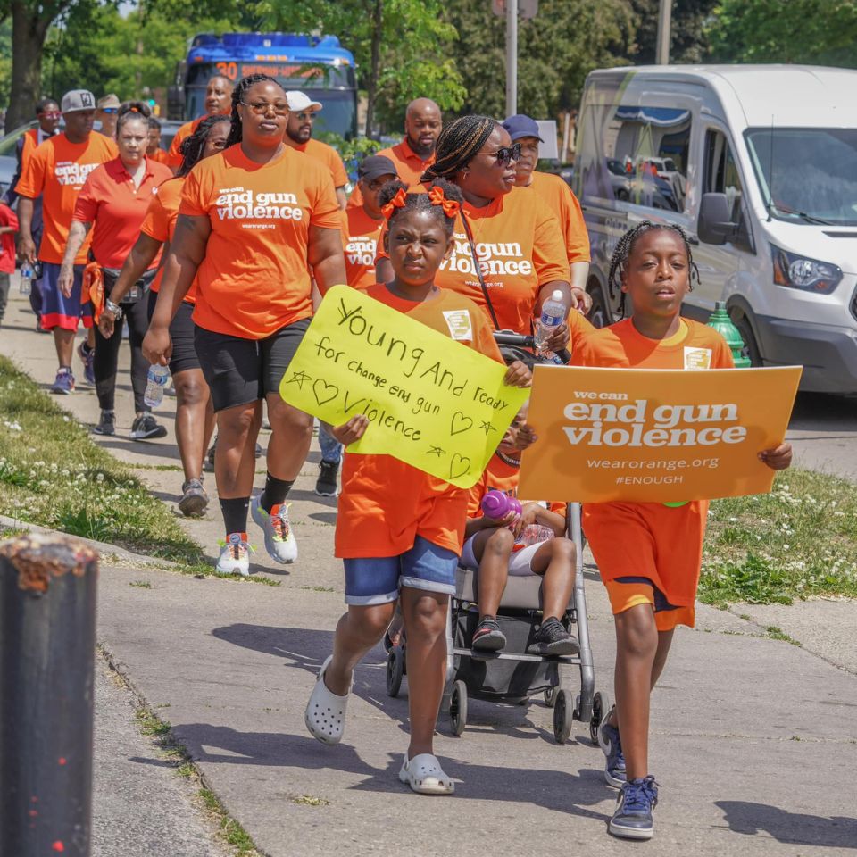 A line of marchers walks on a sidewalk. The two people in front are two girls of about 7 years old. They each hold signs that say "end gun violence."