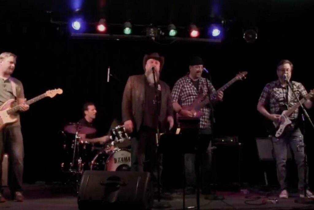 5 musicians are performing. From left to right is a guitarist, drummer, lead singer, bass player and another guitarist. They are wearing plaid shirts and tshirts.