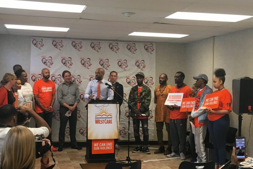 Mandela Barnes speaks at a podium decorated with a WestCare poster and a poster that says We can end gun violence. People stand around and behind him wearing orange. In the foreground, cameras are recording Barnes and snapping photos.