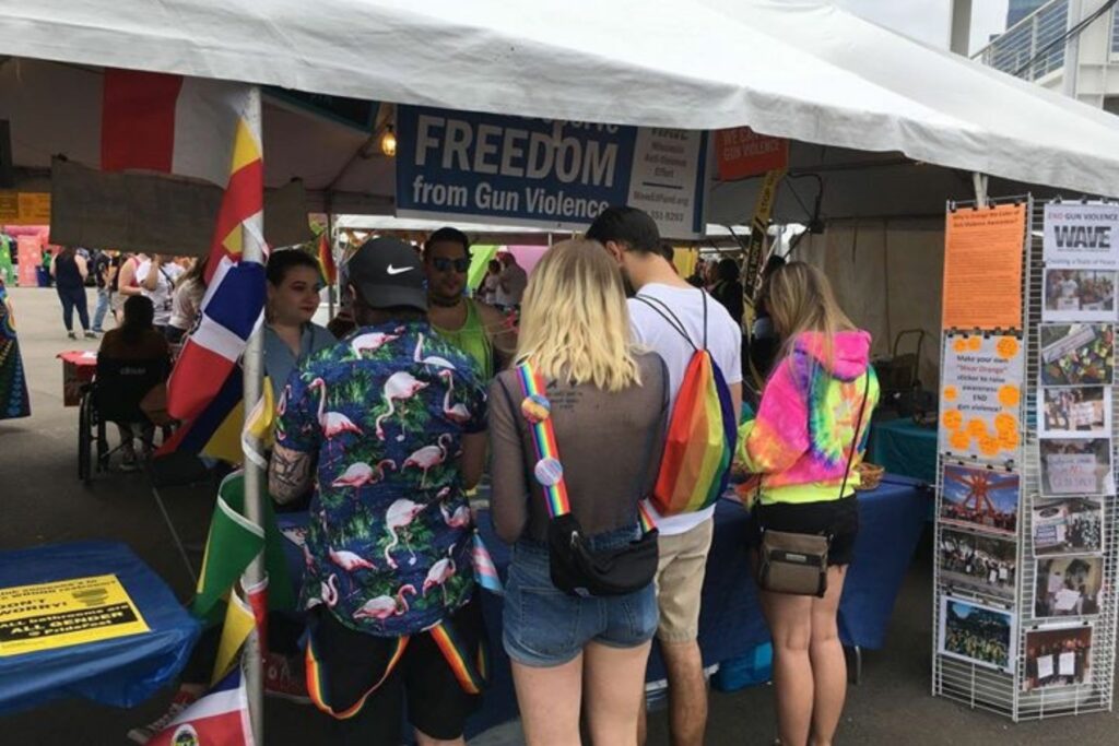 several people are gathered around WAVE's resource table at Pridefest. They wear rainbow bags and tye-die tshirts.