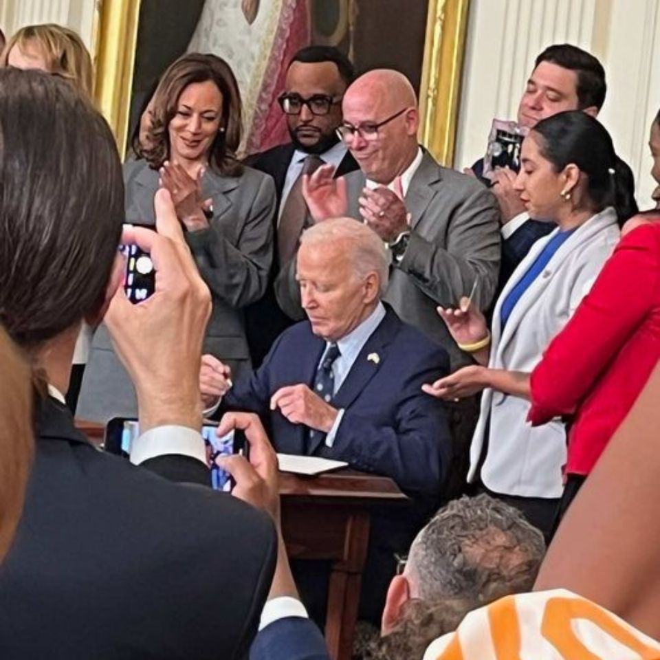 President Biden sits at a table. Behind him stands Vice President Harris, and several survivors of gun violence. The president is signing an executive order to help with gun violence prevention work.