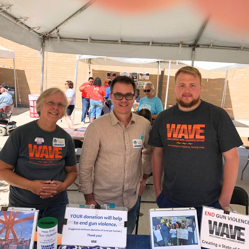 Two WAVE staff and a volunteer stand behind a table at an event. The table is covered in information about gun violence prevention and WAVE.