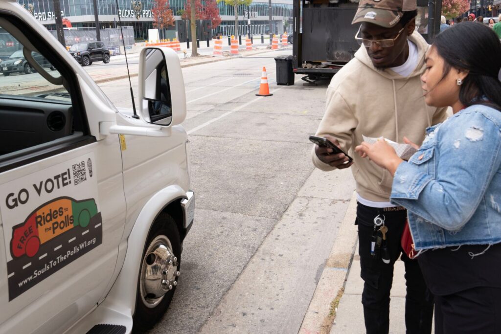 Two people look at a phone together, figuring out logistics. They are next to a van that says Go Vote! Rides to the Polls! It is clear that one of them is getting ready to head to their poling place.