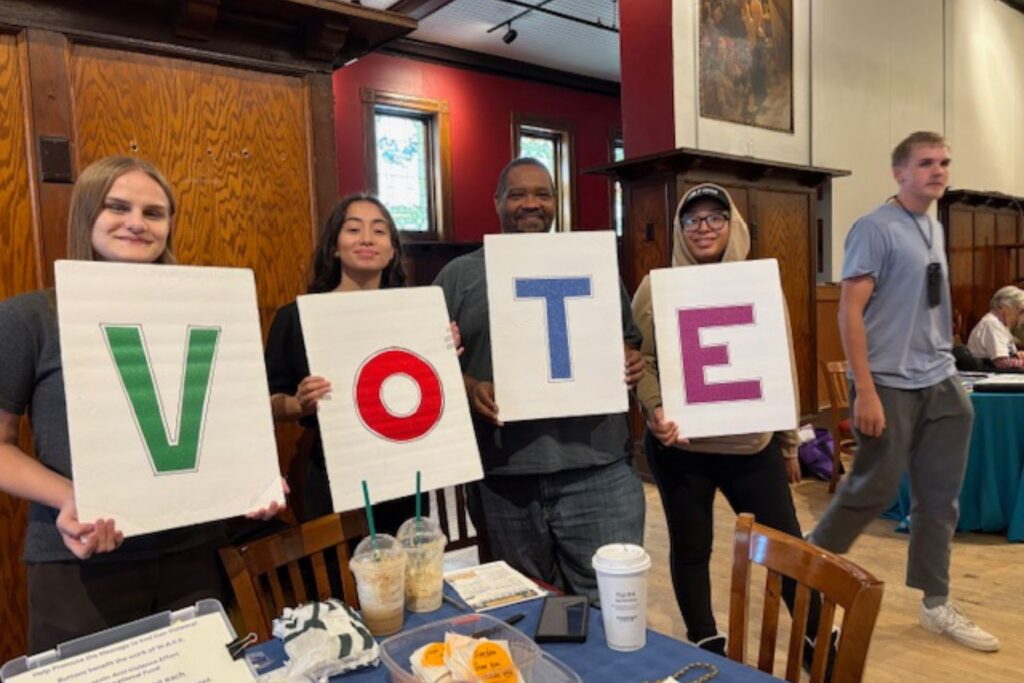 Four people stand in a row, each holding a sign with one letter. Together the letters spell VOTE.