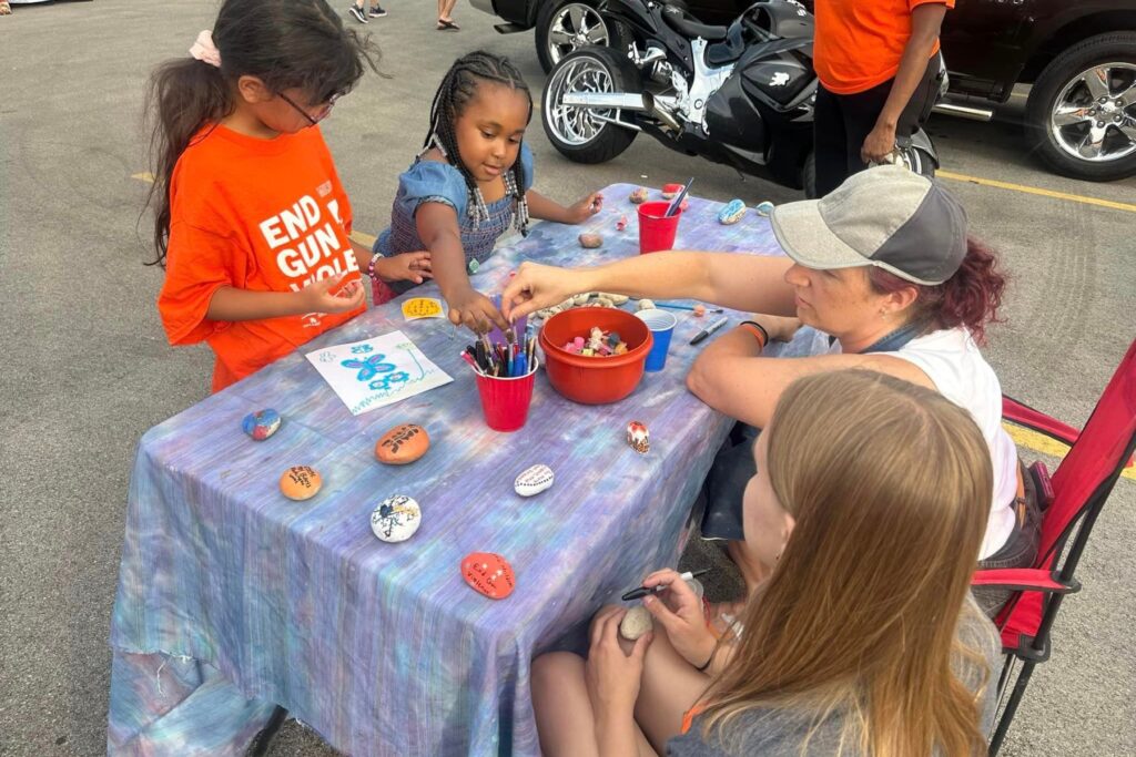 A table with art supplies and painted rocks has people gahered around. Two young children are working on drawing or painting rocks.