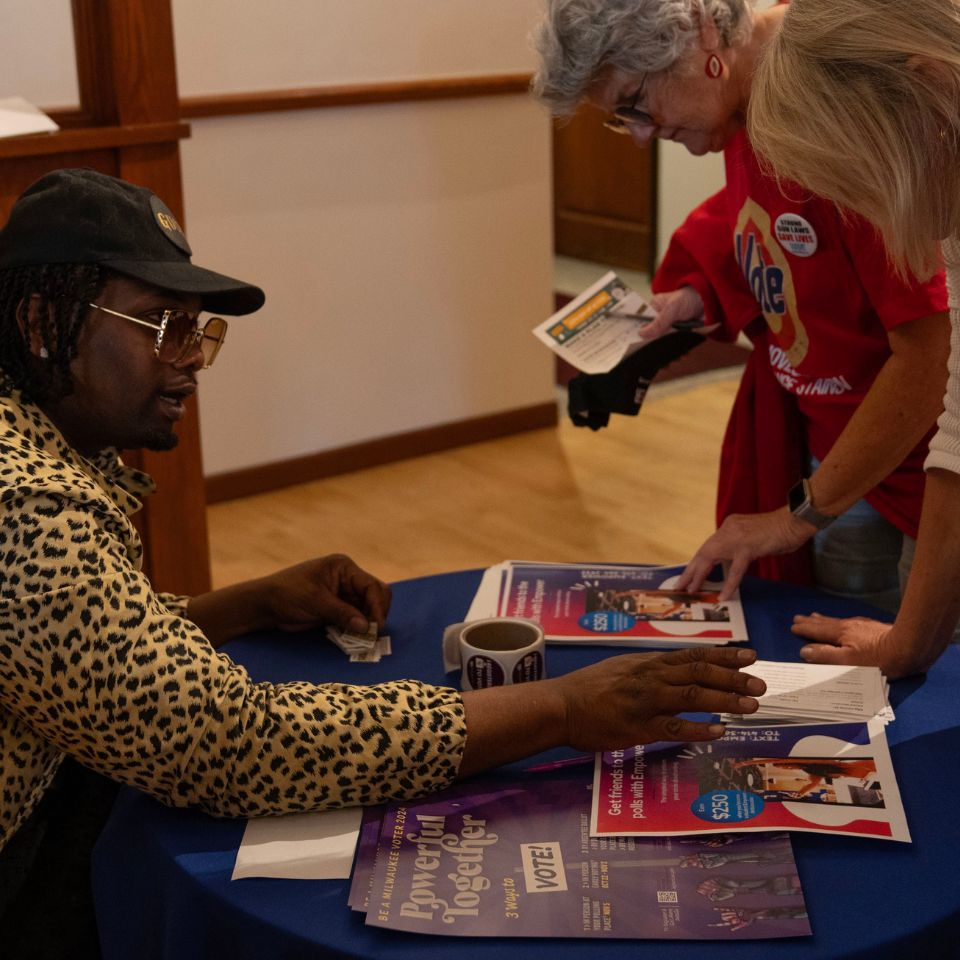 A person wearing leopard print and shiny glasses sits at a table. They are talking with two other people, explaining something as the others bend over looking at information.