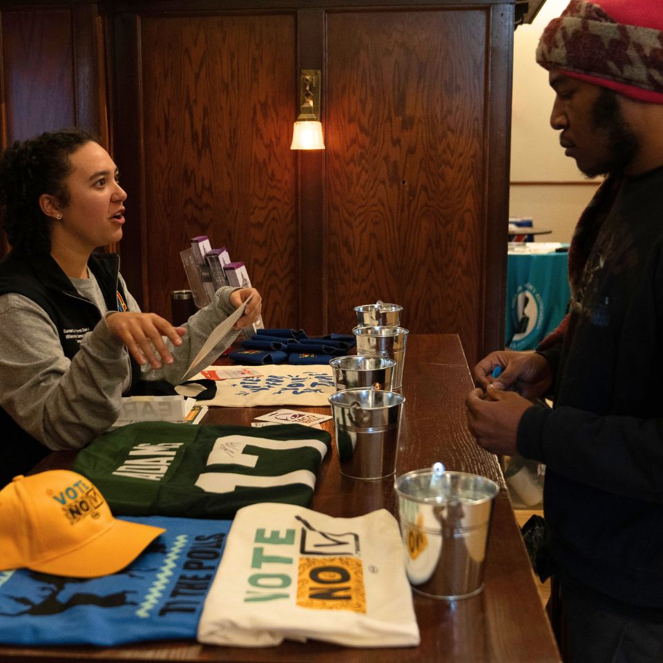 A person stands behind a counter covered in merchandise that says "Vote No." Someone is standing on the other side of the counter, looking at the objects.