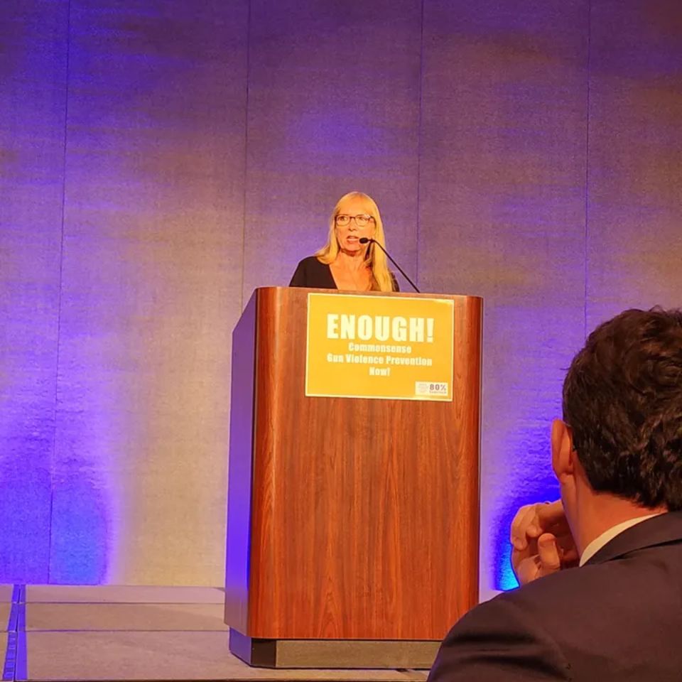 WAVE's Executive Director, Jeri Bonavia, stands on a stage speaking. She is behind a podium, and the podium has a sign on it that says Enough! Commonsense gun violence prevention now!