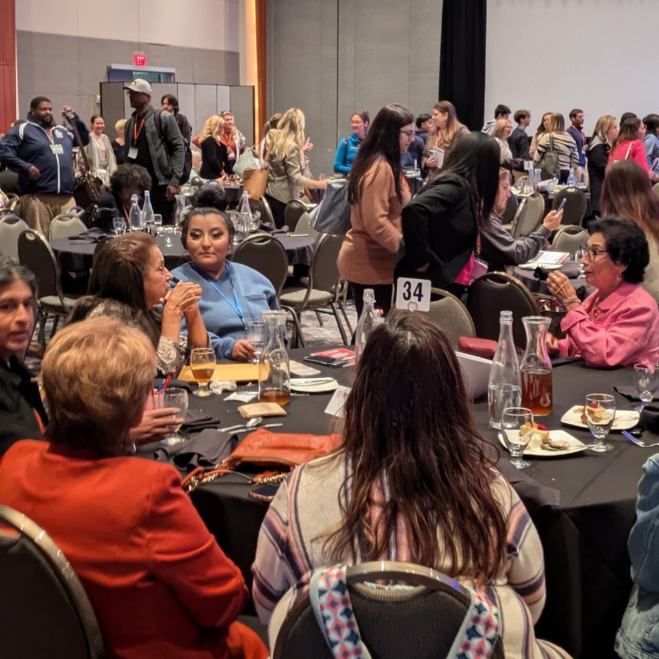 Many people mill around a room during the Gun Violence Summit. Some are sitting, some standing, people are engaged and talking with each other.