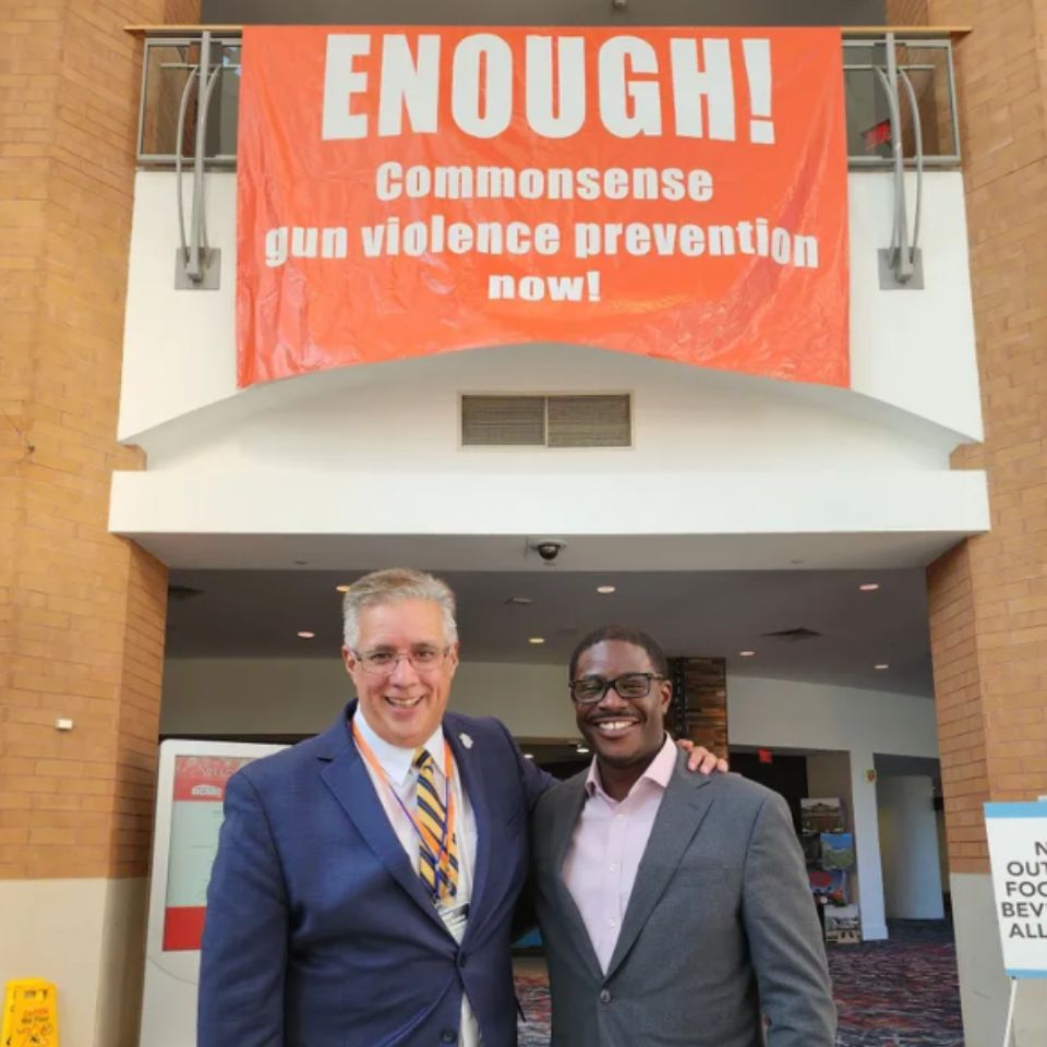 Two people stand beneath a large orange banner that says Enough! Commonsense gun violence prevention now!