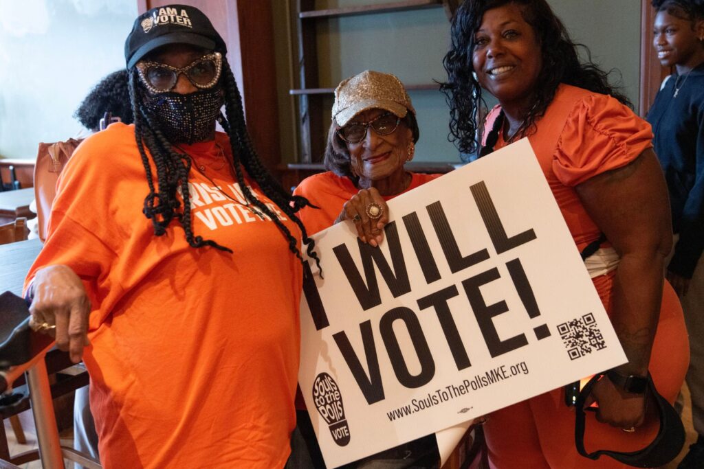 Three people stand together looking at the camera. They wear orange and hold a sign that says I will vote!