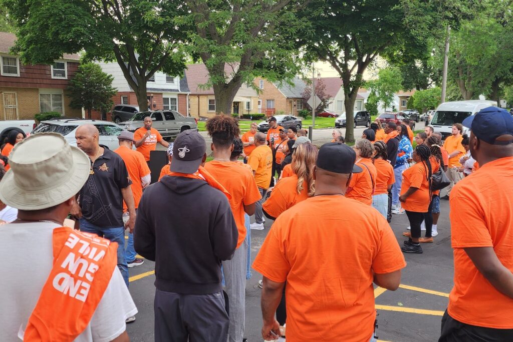 During Milwaukee's Wear Orange events, a speaker stands outside holding a microphone. The foreground of the photo shows dozens of people gathered around, listening.